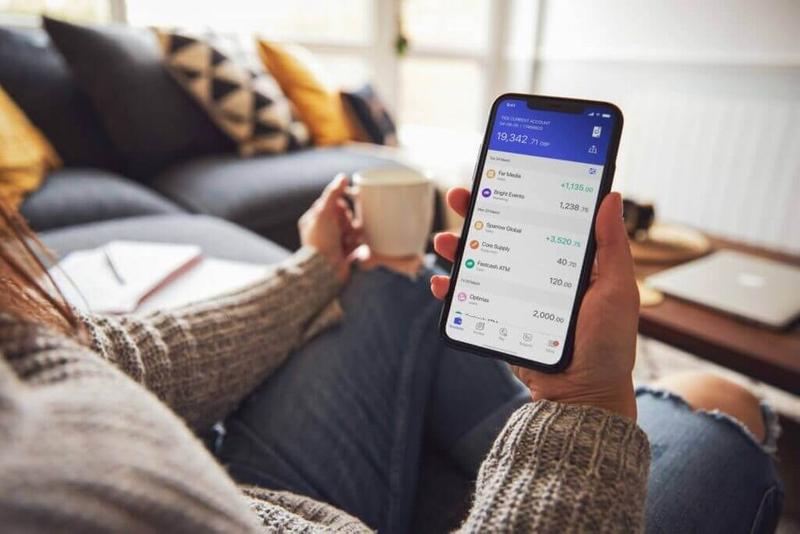 A woman sits on the sofa drinking a coffee and looking at her Tide account on her mobile phone.